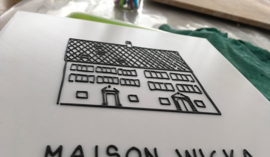 Plaque blanche avec un dessin d'une maison et le texte "MAISON WICKA" en dessous.