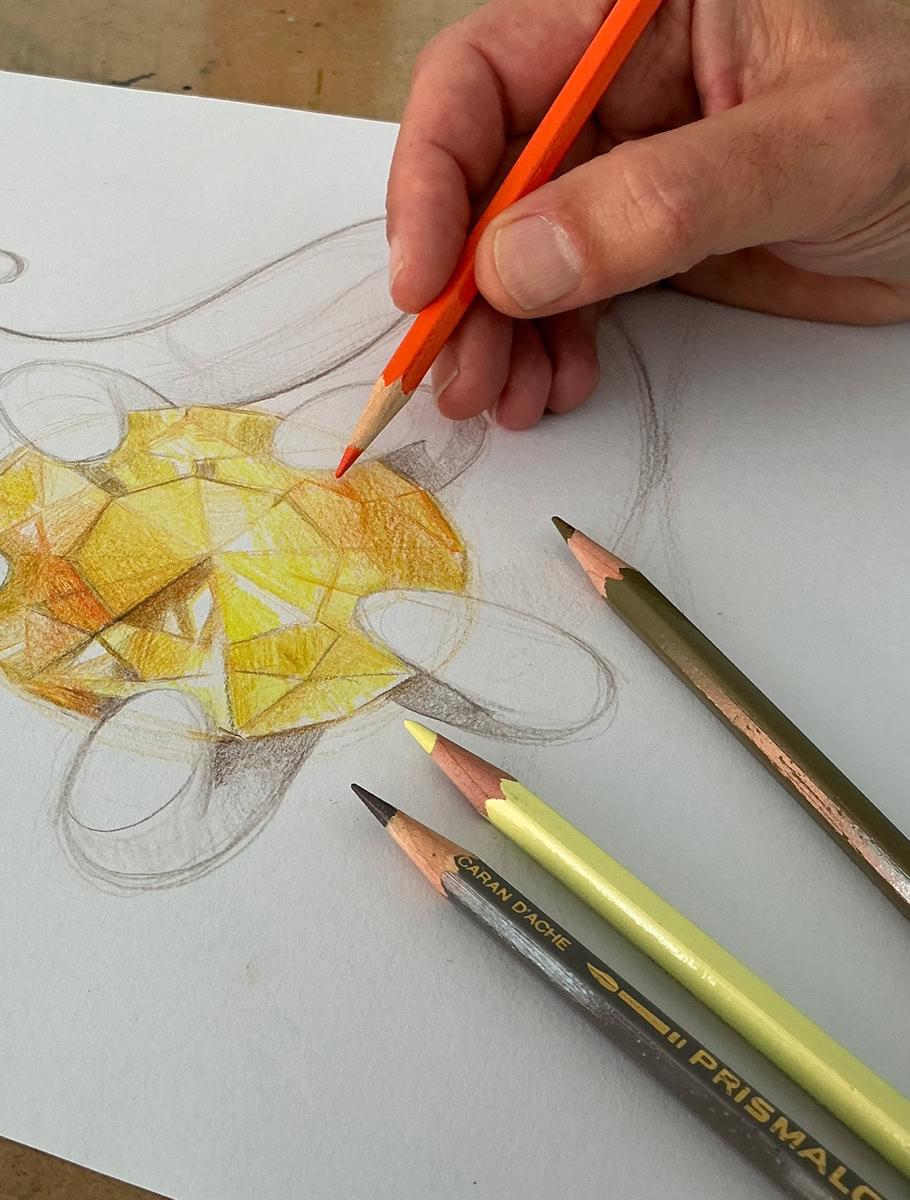 Une main colorie un dessin de  bijou avec une pierre précieuse avec des crayons de couleur, dont un jaune et un orange.