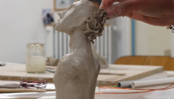 Une main sculpte une tête en argile sur une table, avec des outils et des pots en arrière-plan.