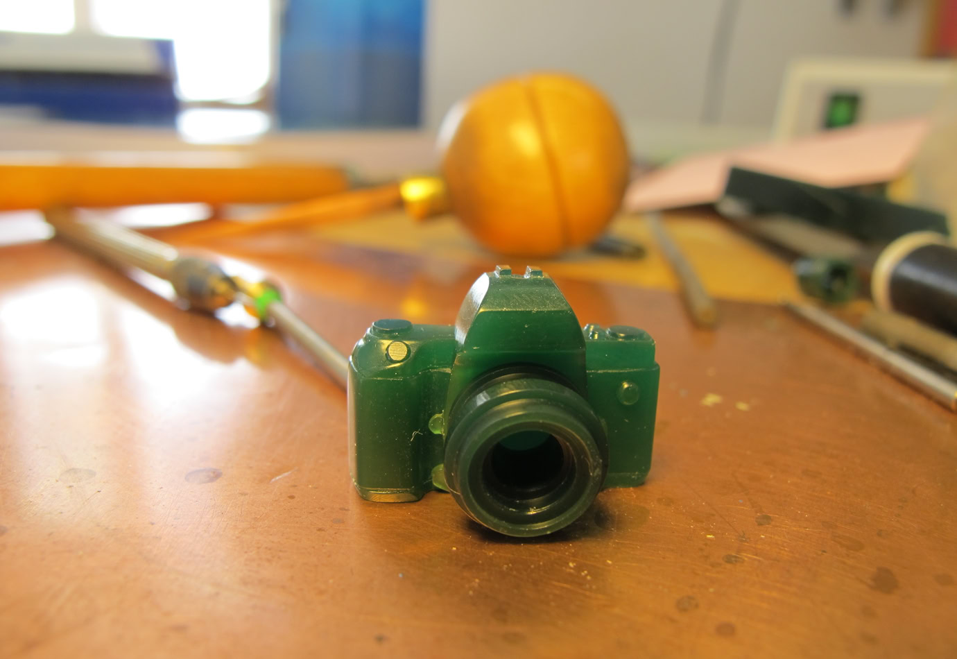 Petite sculpture verte en forme d'appareil photo sur une surface en bois, avec des outils flous en arrière-plan.