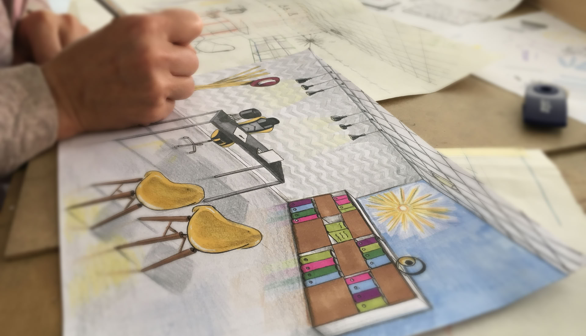 Une main dessine sur une feuille avec un croquis coloré d'une pièce, comprenant des chaises jaunes, une étagère avec des livres et un mur bleu avec un soleil dessiné.