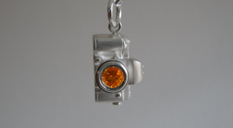 Pendentif en forme d'appareil photo argenté avec un détail orange, suspendu à une chaîne.