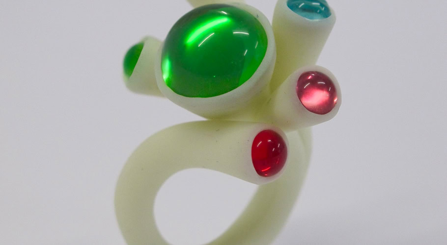 Un crayon rose traversant une bague en résine blanc avec des embouts colorés, entouré de crayons de couleur sur une table.