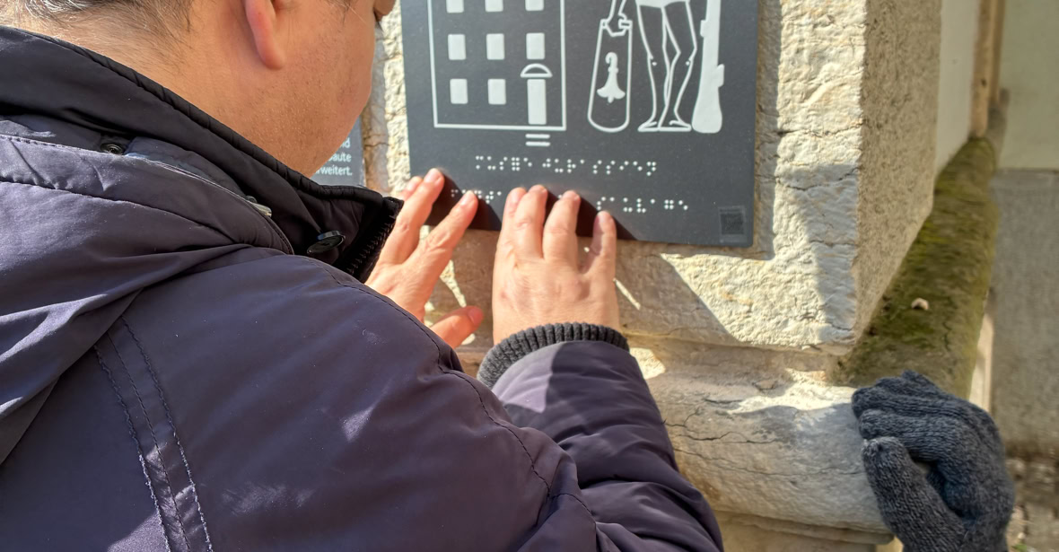 Deux personnes lisant un dessin tactil sur une plaque sur un mur en pierre, avec un dessin d'un bâtiment et d'une figure humaine.