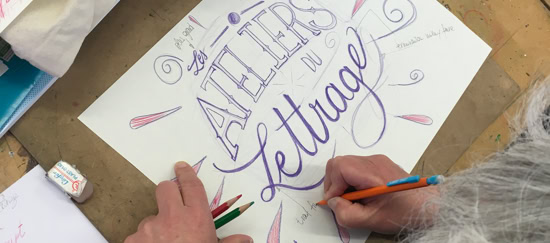 Personne faisant de la calligraphie sur une feuille de papier avec des crayons, avec les mots "ATELIERS de lettrage" visibles.