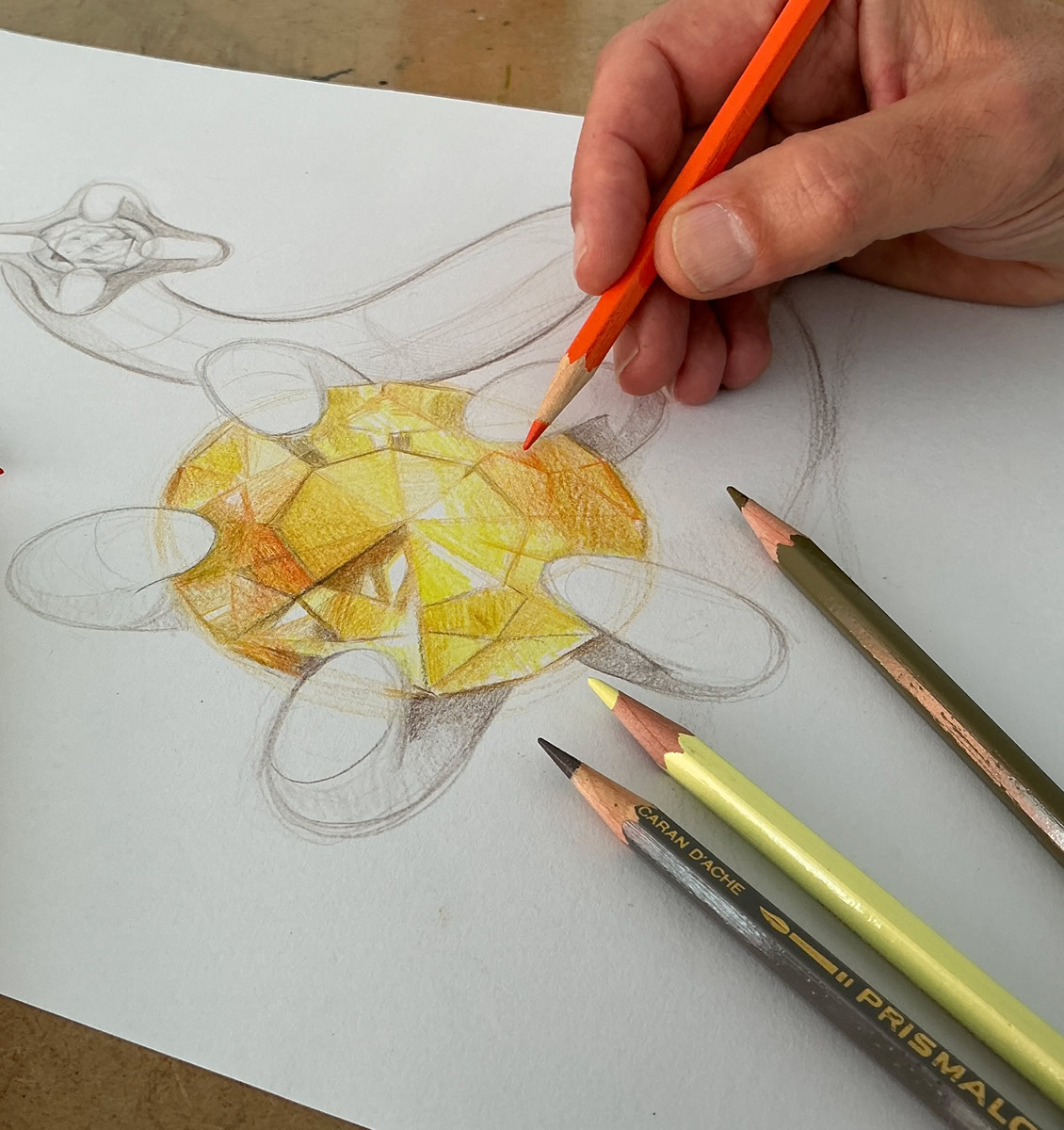 Une main colorie un dessin de  bijou avec une pierre précieuse avec des crayons de couleur, dont un jaune et un orange.