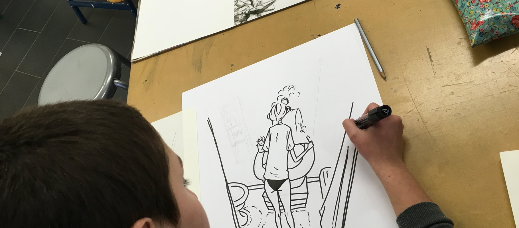 Un enfant dessine une illustration en noir et blanc sur une feuille de papier, avec d'autres feuilles et un étui à crayons sur la table.