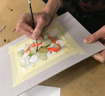 Personne peignant une aquarelle sur une feuille de papier fixée avec du ruban adhésif, pinceaux à proximité.