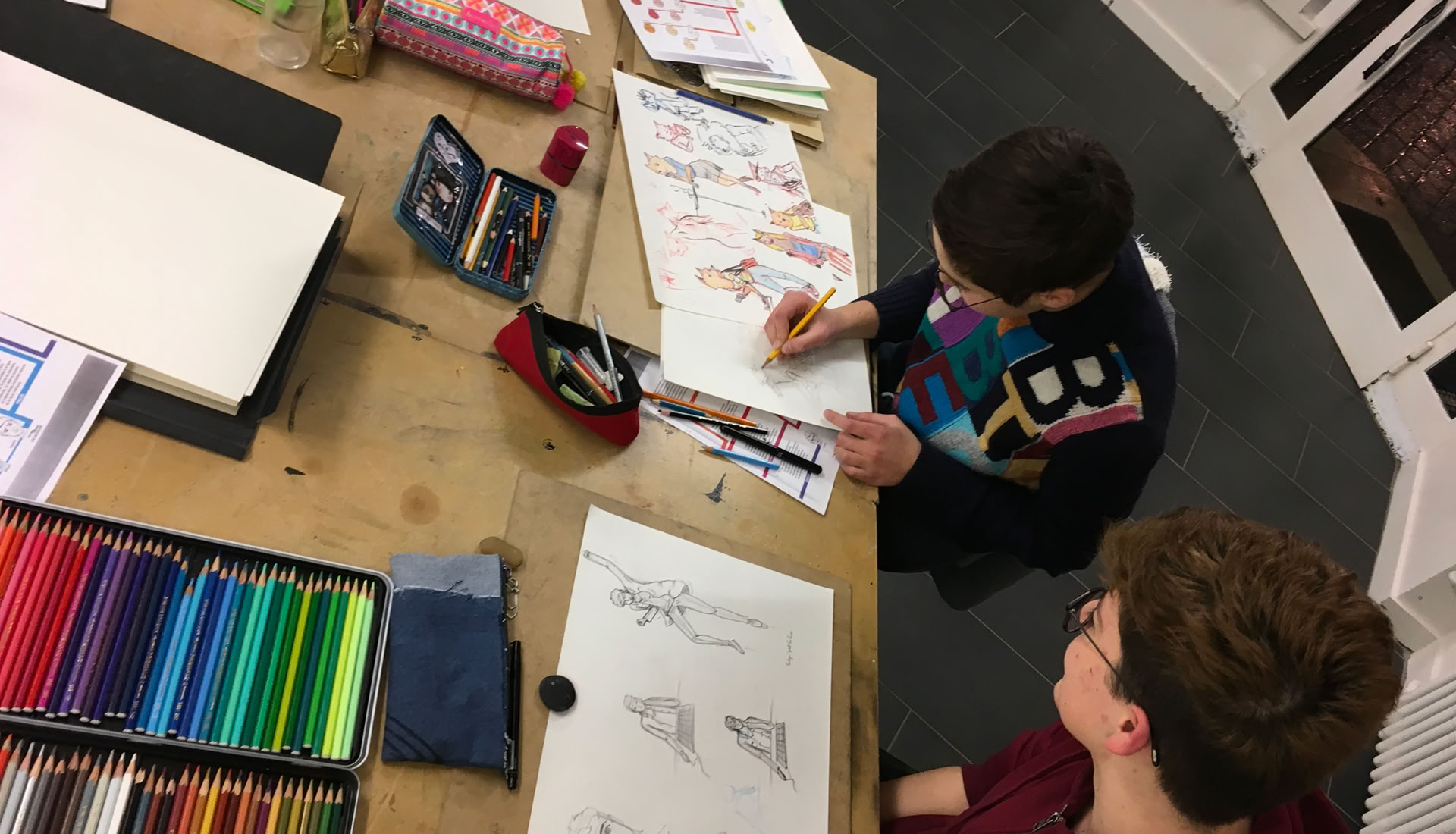Deux personnes assises à une table en train de dessiner, avec des crayons de couleur, des carnets de croquis et d'autres fournitures artistiques dispersés sur la table.