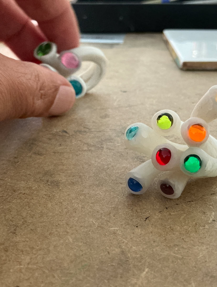Main tenant une bague en résine blanc avec des perles colorés, posé sur une surface en bois.