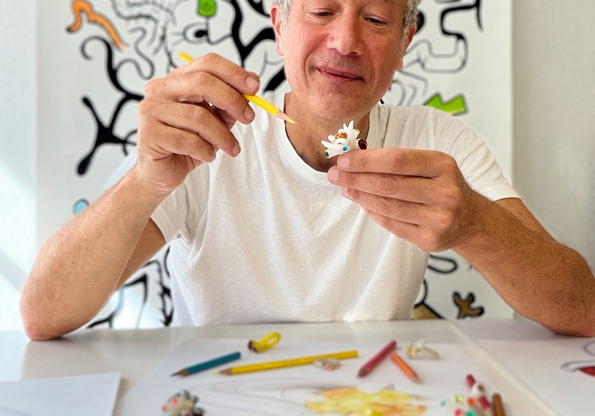 Un homme assis à une table avec des crayons de couleur et du papier, tenant un pinceau et une petite sculpture. En arrière-plan, un tableau avec des motifs abstraits.