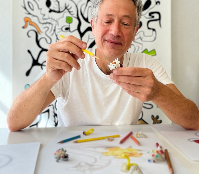 Un homme assis à une table avec des crayons de couleur et du papier, tenant un pinceau et une petite sculpture. En arrière-plan, un tableau avec des motifs abstraits.