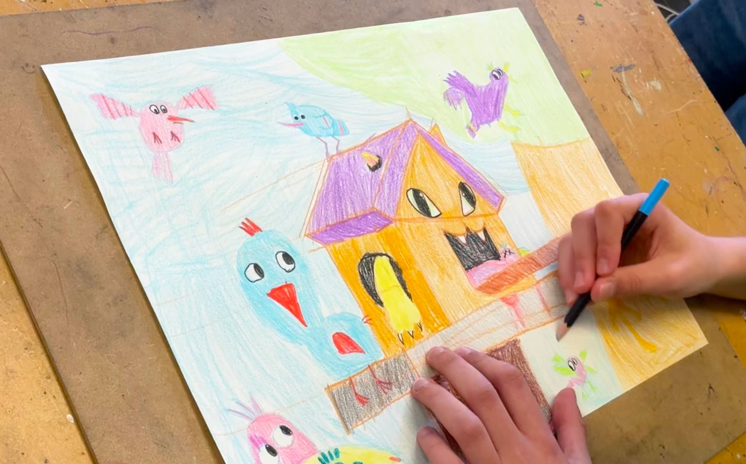 Une personne dessine sur une feuille de papier avec des crayons de couleur, représentant une maison colorée entourée d'oiseaux.