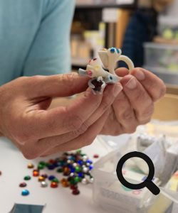Deux mains tiennent une petite sculpture en résine avec des pierres en verre de Bohème. Des perles colorées et une boîte de rangement sont visibles sur la table.