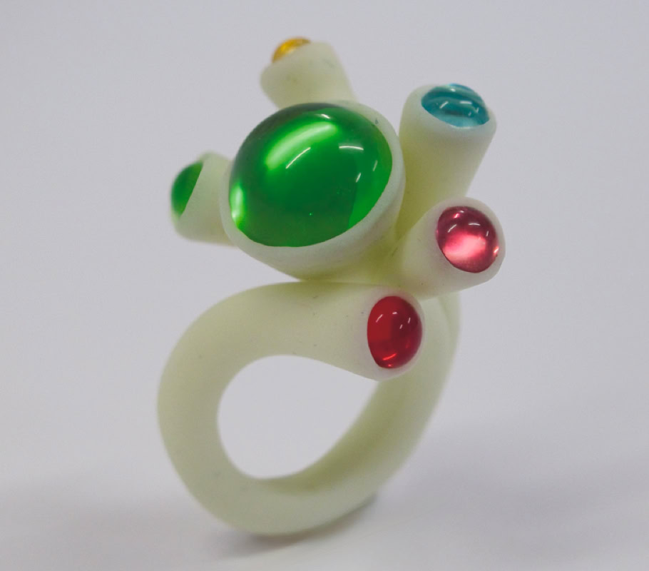 Bague blanche avec des pierres vertes, rouges et bleues sur un fond blanc.