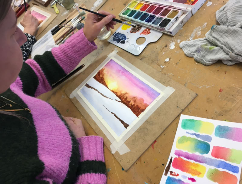 Une personne peint une aquarelle représentant un paysage avec des arbres et un ciel coloré. Des palettes de peinture et des pinceaux sont visibles sur la table.