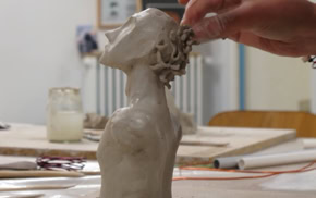 Une main sculpte une tête en argile sur une table, avec des outils et des pots en arrière-plan.