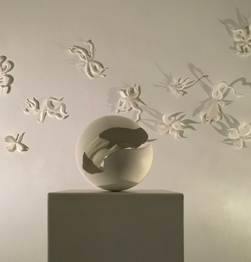 Sculpture en forme de sphère sur un socle, entourée de papillons blancs en relief sur un mur.
