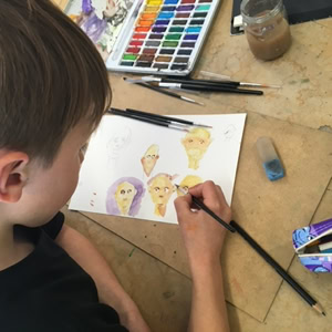 Enfant dessinant sur du papier avec des crayons et des aquarelles, boîte de crayons et gomme à côté.