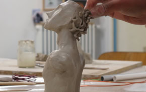 Une main sculpte une tête en argile sur une table, avec des outils et des pots en arrière-plan.