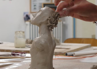 Une main sculpte une tête en argile sur une table, avec des outils et des pots en arrière-plan.