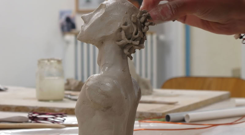 Une main sculpte une tête en argile sur une table, avec des outils et des pots en arrière-plan.