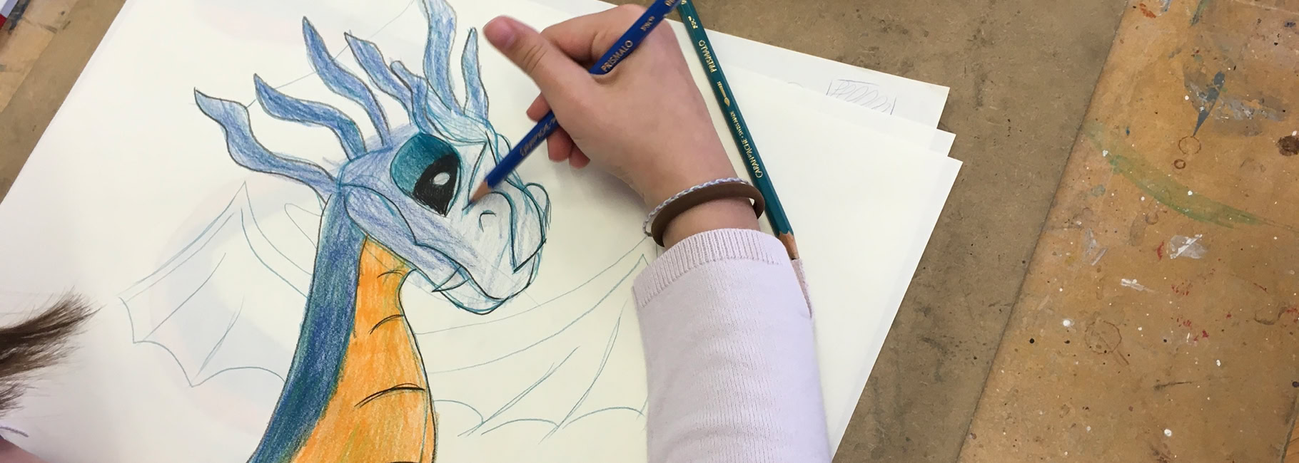 Une personne dessine un dragon coloré sur du papier avec des crayons de couleur, une trousse rose est visible en arrière-plan.