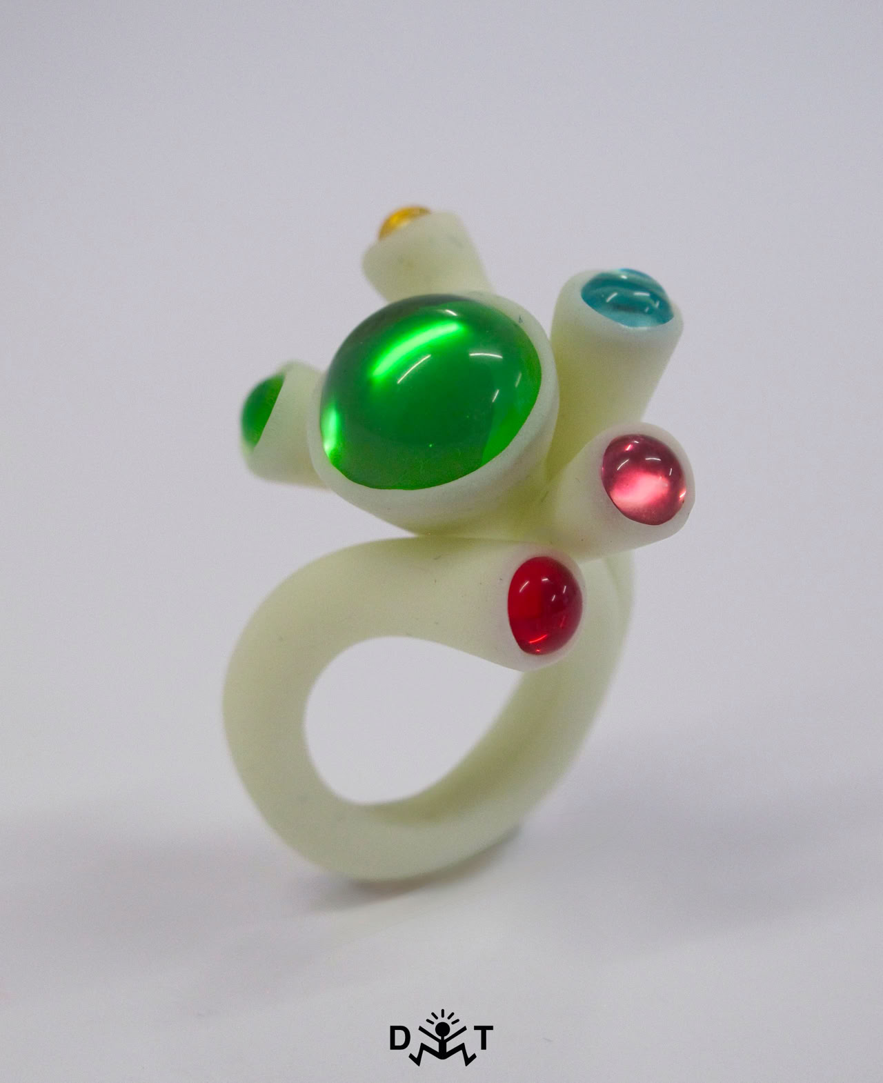 Bague blanche avec des pierres colorées vertes, rouges et bleues sur un fond blanc.