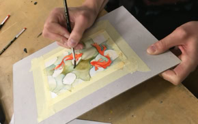 Personne peignant une aquarelle sur une feuille de papier fixée avec du ruban adhésif, pinceaux à proximité.