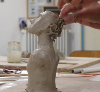 Une main sculpte une tête en argile sur une table, avec des outils et des pots en arrière-plan.