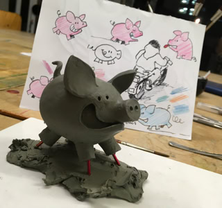 Sculpture en argile d'un cochon souriant sur une table, avec un dessin de cochons et une personne en arrière-plan.