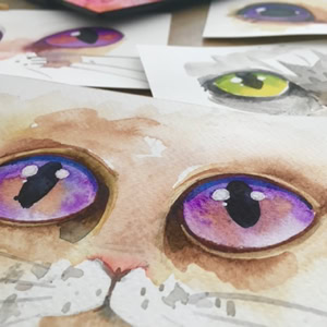Peintures aquarelles de visages de chats avec de grands yeux colorés, principalement violets et verts, disposées sur une surface.
