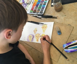 Enfant dessinant sur du papier avec des crayons et des aquarelles, boîte de crayons et gomme à côté.