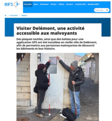 Deux personnes debout près d'un mur, l'une tenant une canne blanche et l'autre pointant vers un panneau.