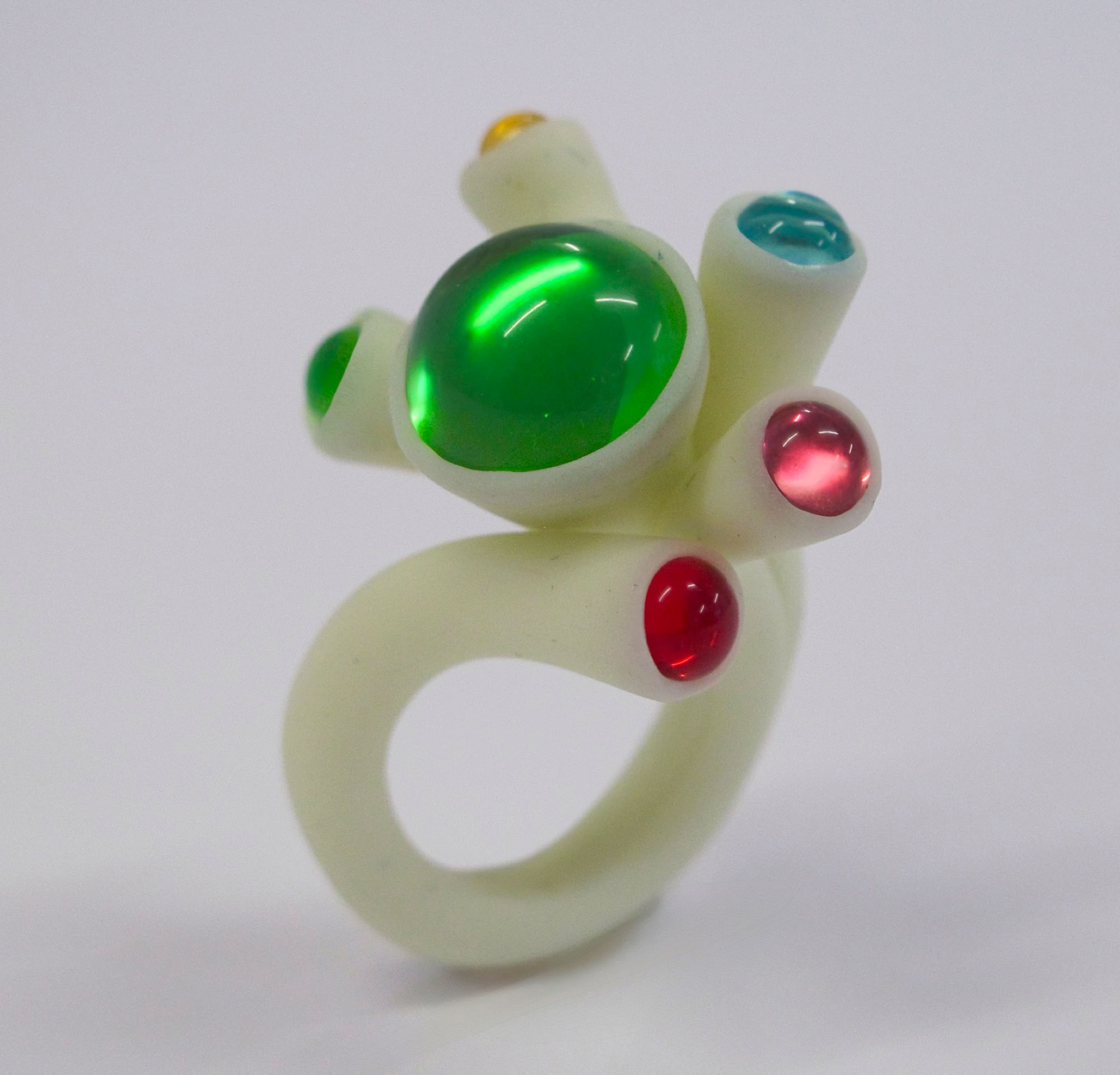 Bague blanche avec des pierres colorées vertes, rouges et bleues sur un fond blanc.