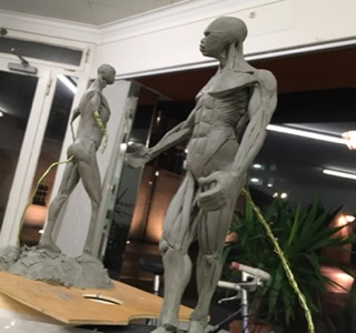 Deux sculptures en argile représentant des figures humaines musclées dans un atelier, avec des plantes en arrière-plan.