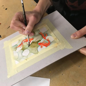 Personne peignant une aquarelle sur une feuille de papier fixée à une planche, avec des pinceaux à proximité.