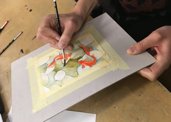 Personne peignant une aquarelle sur une feuille de papier fixée avec du ruban adhésif, pinceaux à proximité.