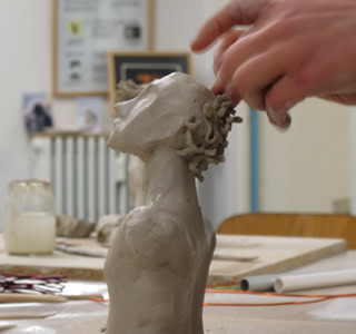 Une main sculpte une petite statue en argile représentant une tête humaine sur une table, avec des outils et des objets en arrière-plan.