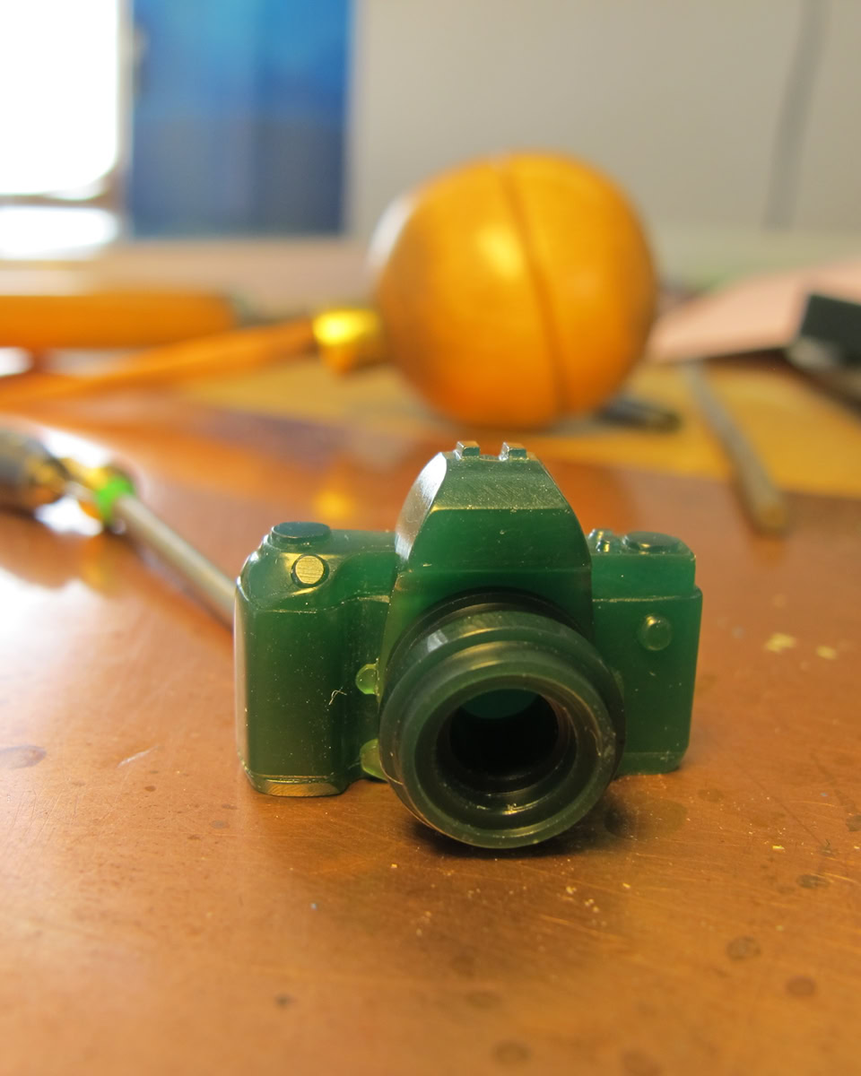 Petite sculpture verte en forme d'appareil photo sur une surface en bois, avec des outils flous en arrière-plan.
