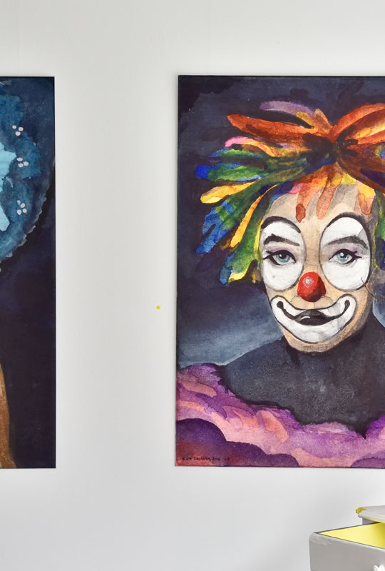 Deux peintures accrochées au mur, l'une représentant une personne avec des cheveux bleus et l'autre un clown avec des cheveux multicolores. Sur une table en dessous, des objets décoratifs et une plante.