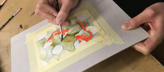Personne peignant une aquarelle sur une feuille de papier fixée avec du ruban adhésif, pinceaux à proximité.