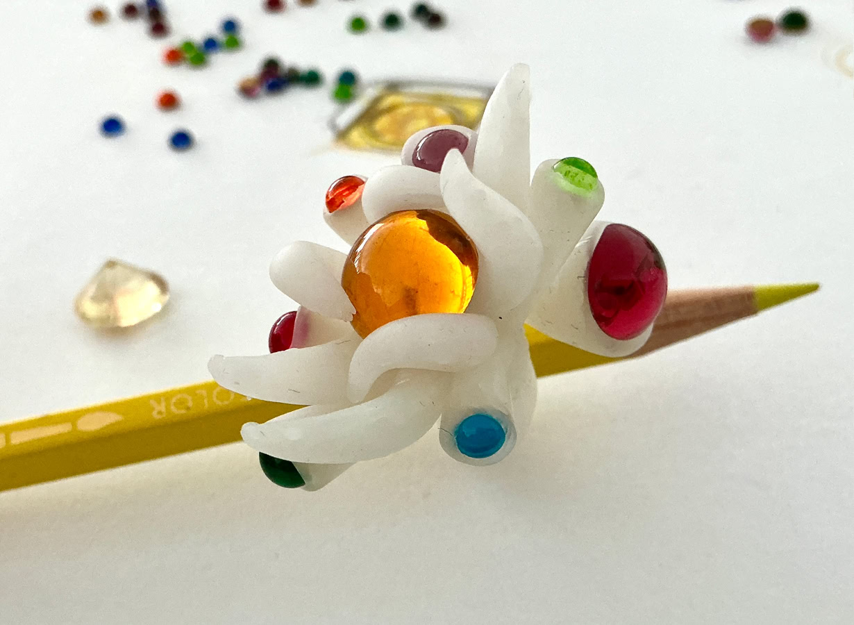 Un crayon jaune avec une bague en résine en forme de fleur blanche ornée de perles colorées, posé sur une surface blanche avec des perles dispersées autour.