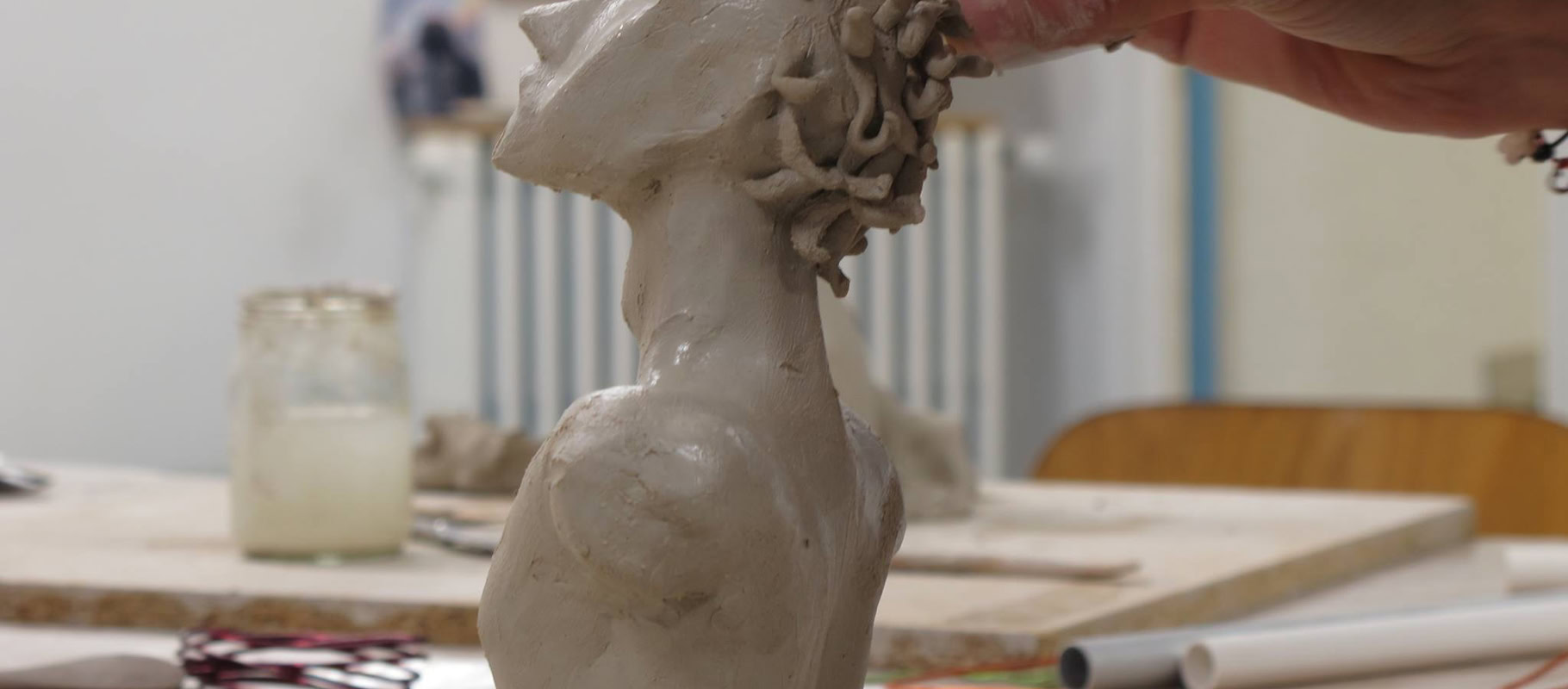 Une main sculpte une tête en argile sur une table, avec des outils et des pots en arrière-plan.