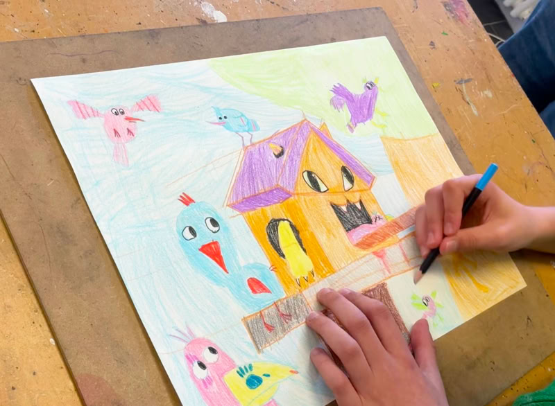 Une personne dessine sur une feuille de papier avec des crayons de couleur, représentant une maison colorée entourée d'oiseaux.
