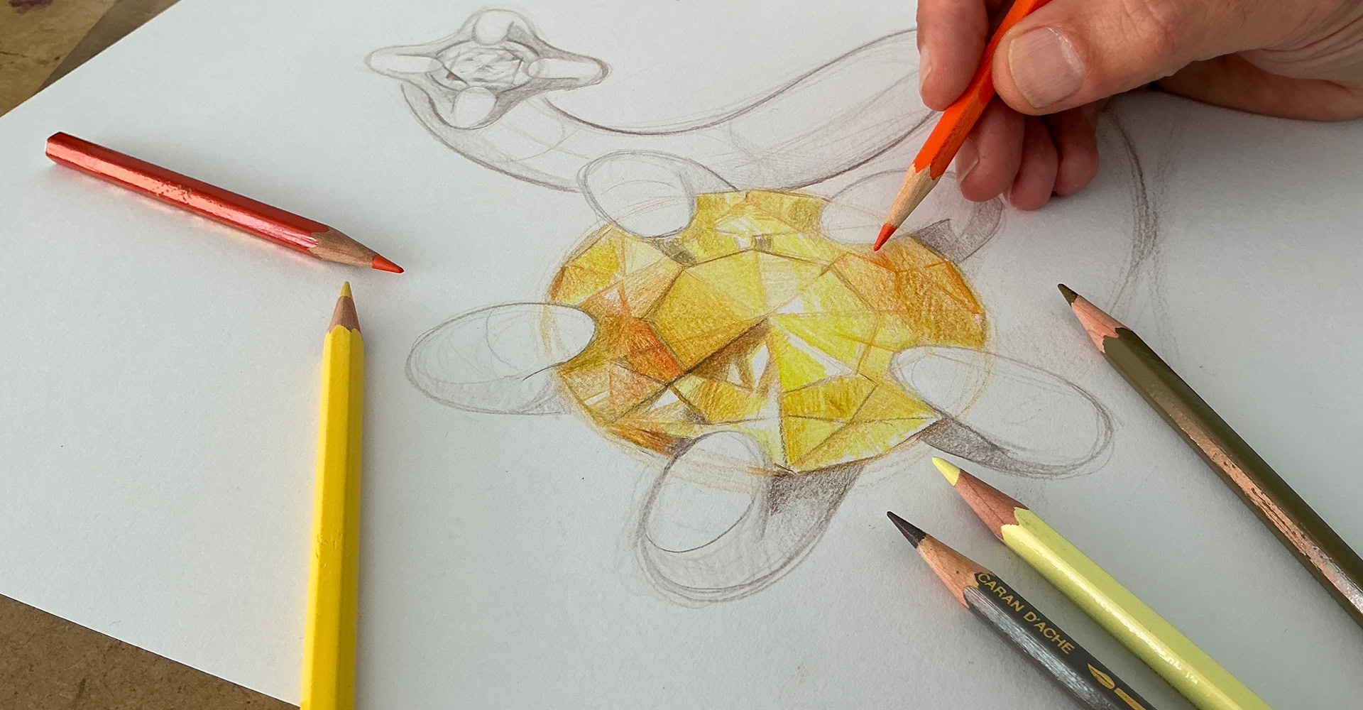 Une main colorie un dessin de  bijou avec une pierre précieuse avec des crayons de couleur, dont un jaune et un orange.