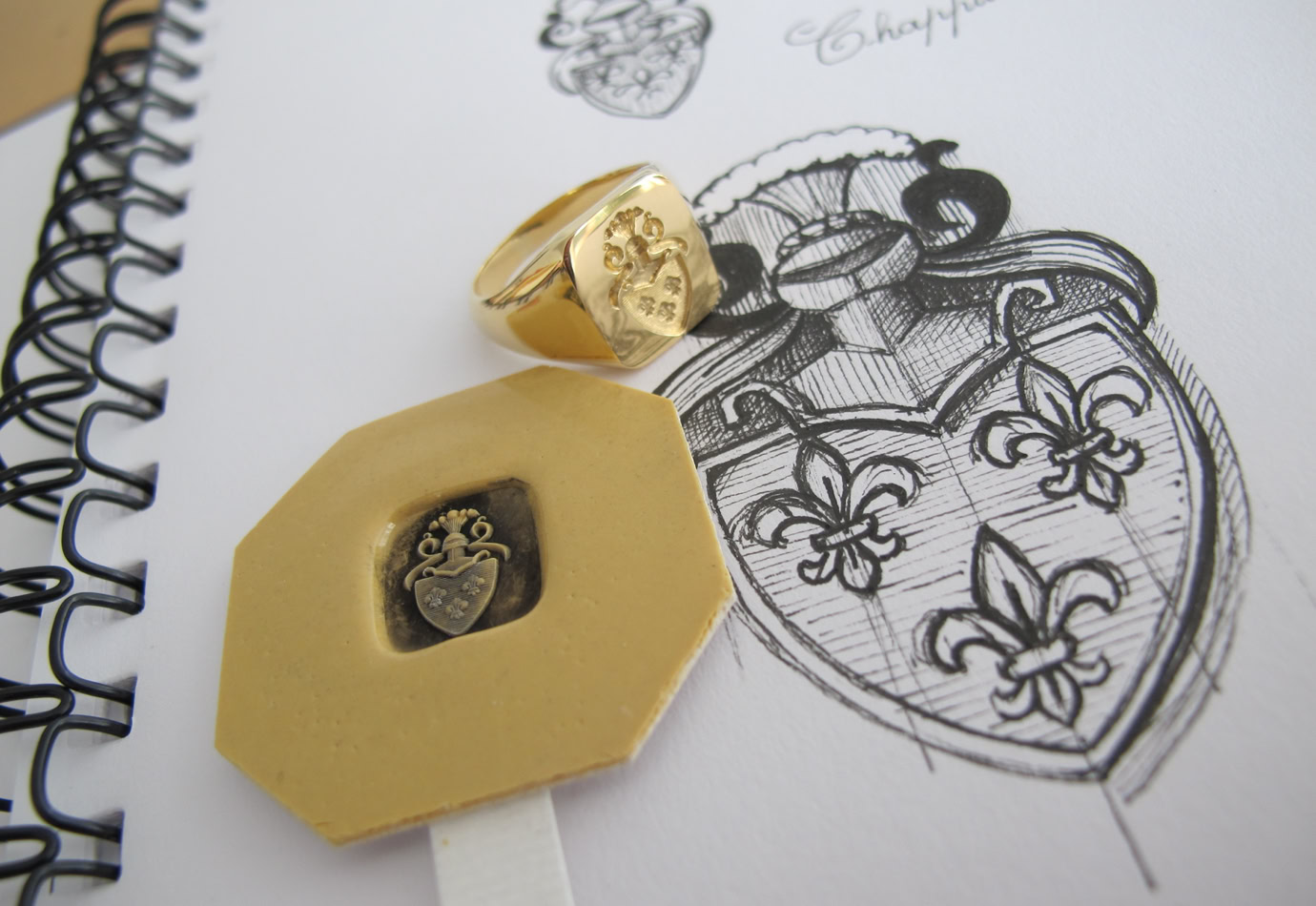 Pièce artistique réalisée sur mesure. Bague Or avec un blason gravé, posée sur un carnet à spirale avec un dessin de blason et un sceau en cire.