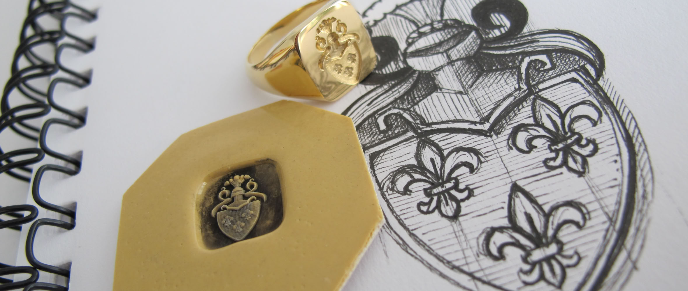 Bague dorée avec un blason gravé, posée sur un carnet à spirale avec un dessin de blason et un sceau en cire.