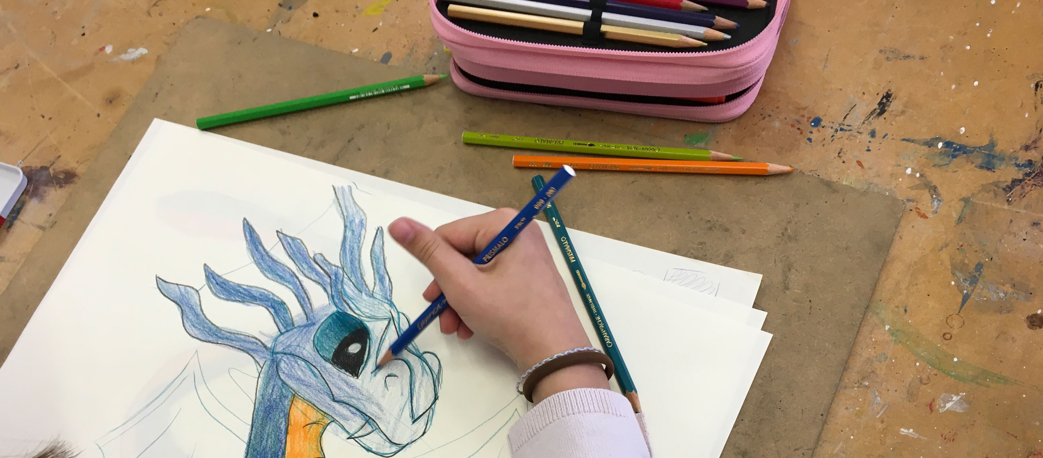 Une personne dessine un dragon coloré sur du papier avec des crayons de couleur, une trousse rose est visible en arrière-plan.