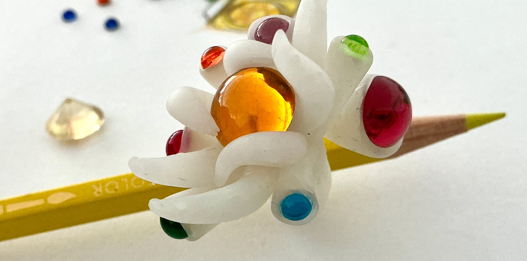 Un crayon jaune avec une bague en résine en forme de fleur blanche ornée de perles colorées, posé sur une surface blanche avec des perles dispersées autour.